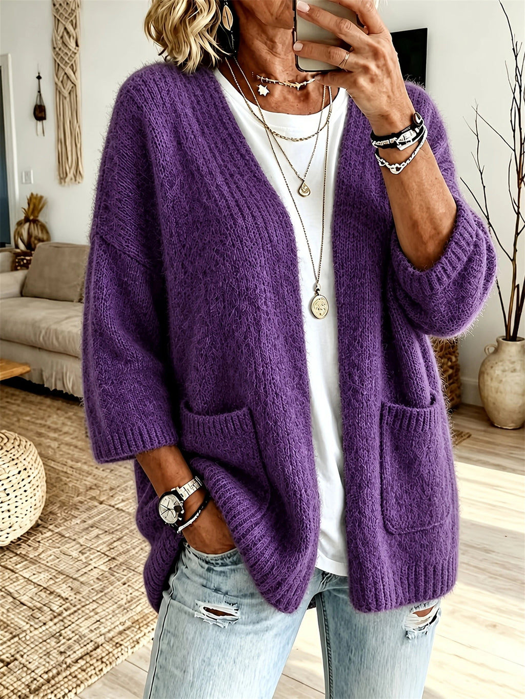 A woman wearing a chunky purple open-front cardigan sweater with front pockets, layered over a white t-shirt and light-wash distressed jeans, in a cozy living room setting.