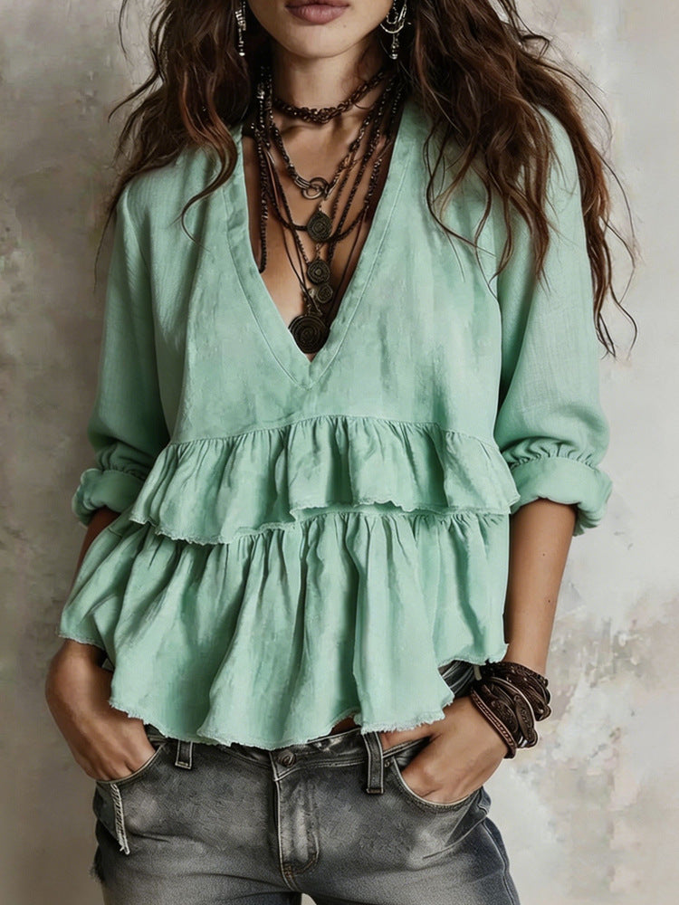 A woman wearing a sage green tiered ruffle blouse with a deep V-neck, styled with layered dark necklaces, bracelets, and gray jeans.