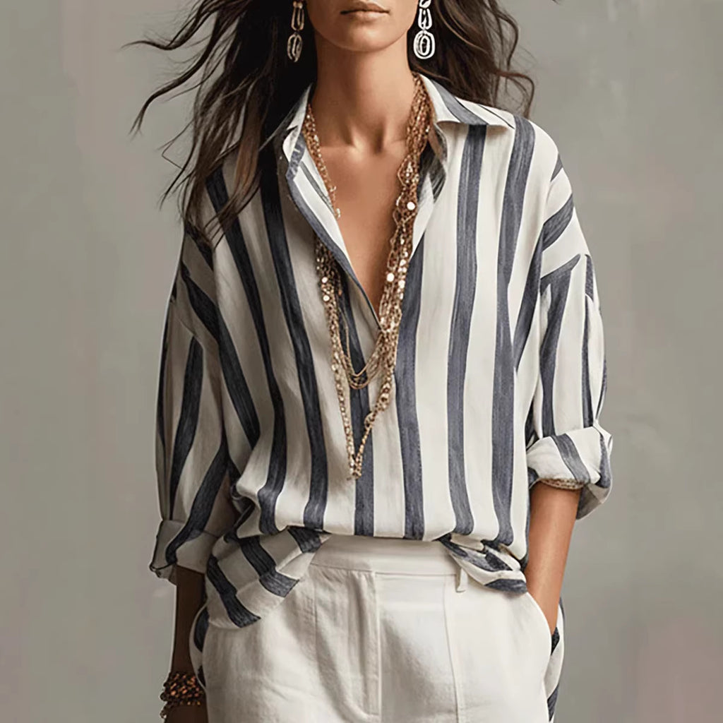 A woman wearing a loose white button-up shirt with vertical navy stripes, styled with layered gold necklaces, white pants, and statement earrings against a soft gray studio background.