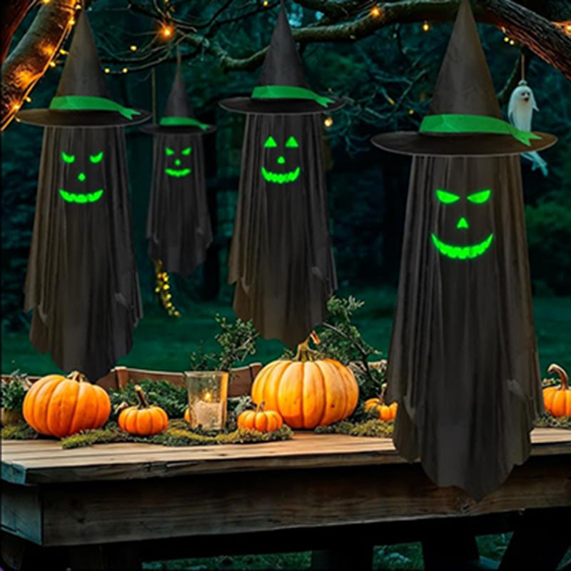 Halloween decorations featuring four hanging witch ghost figures with glowing green faces and pointed black hats, displayed on a wooden table outdoors with pumpkins and string lights.
