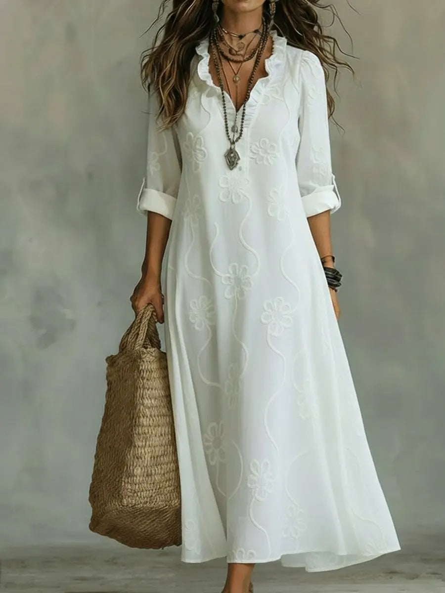 A woman wearing a white bohemian-style midi dress with floral embroidery, holding a woven straw tote bag against a textured gray studio background.