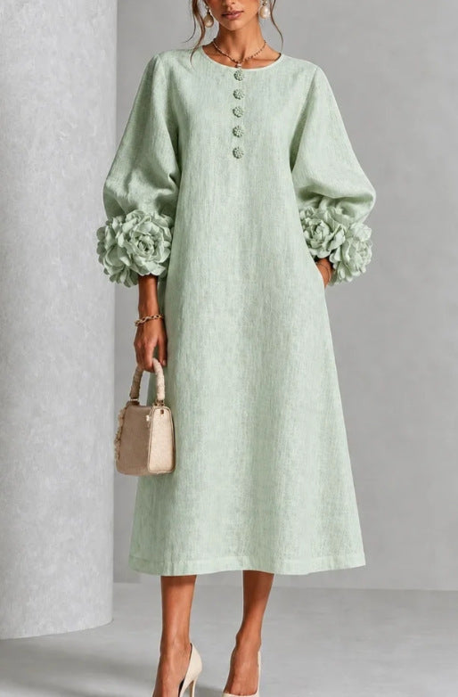 A woman models a light sage green linen midi dress with voluminous long sleeves featuring ruffled cuffs, a round neckline with decorative buttons, and a relaxed A-line silhouette. She is holding a small beige handbag and wearing beige high heels against a neutral gray studio background.