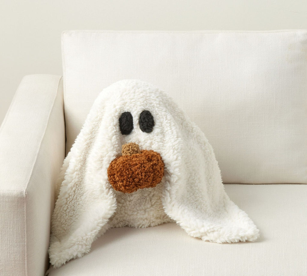 White ghost-shaped pillow with black eyes and a brown pumpkin nose on a white couch.