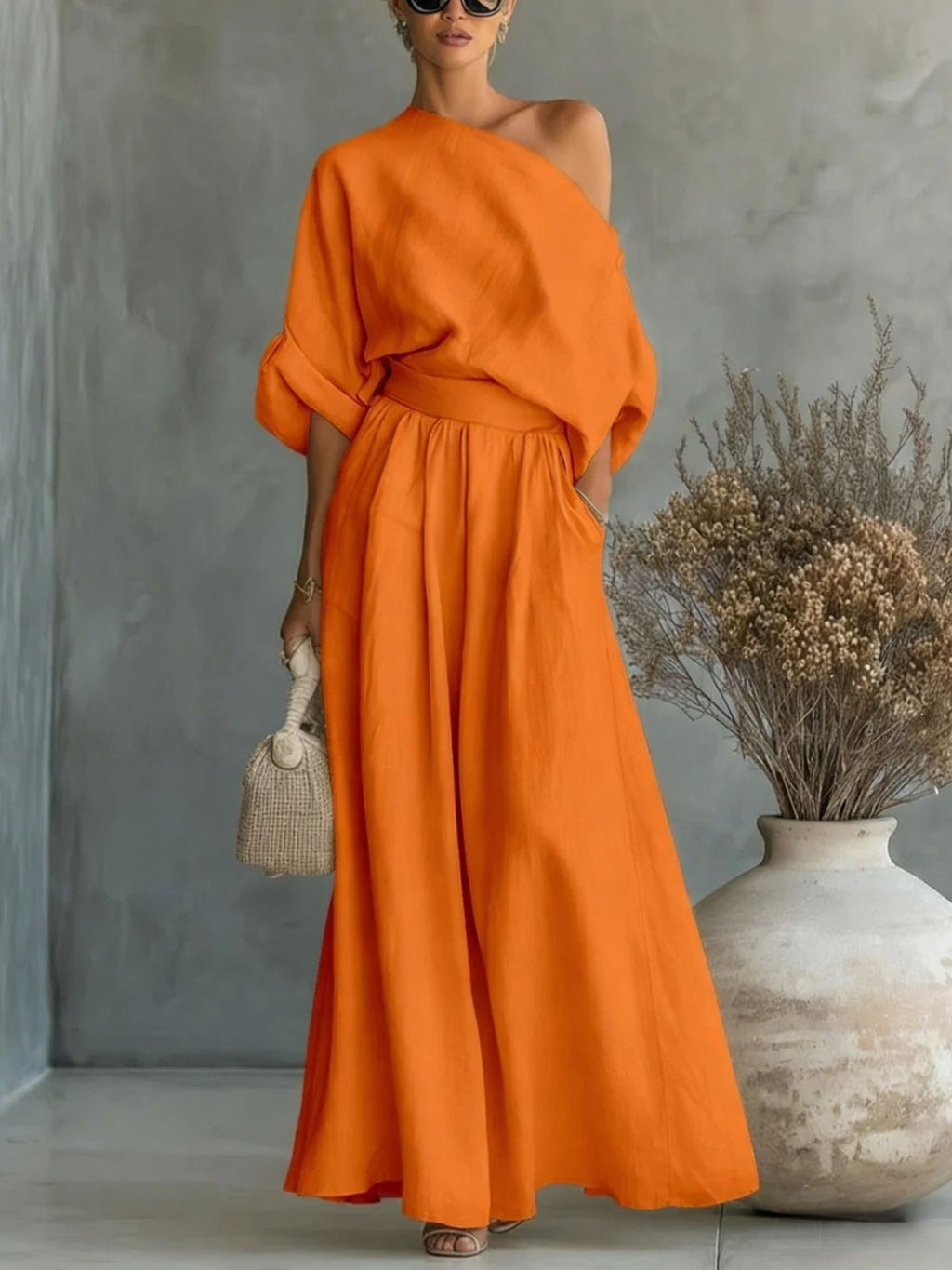 Woman wearing an orange dress standing against a gray wall with a vase and dried plants.