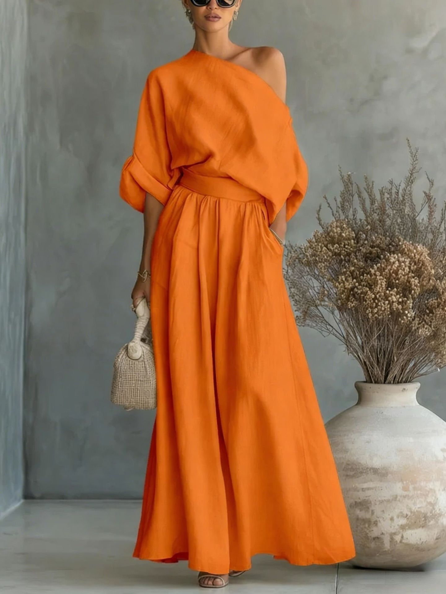 Woman wearing an orange dress standing against a gray wall with a vase and dried plants.