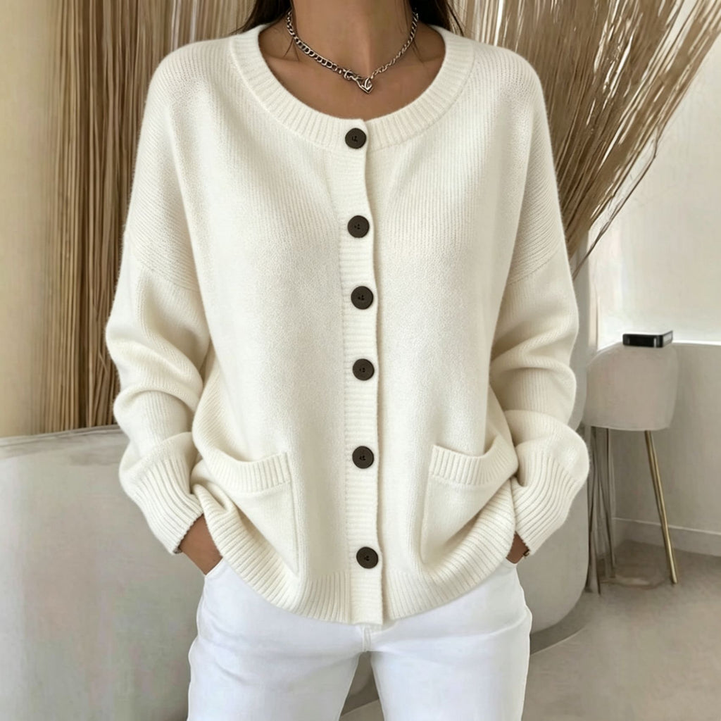 A woman wearing a cream-colored, long-sleeve knit cardigan sweater with dark brown buttons and front patch pockets, paired with white pants.