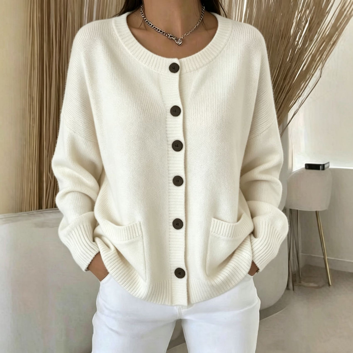 A woman wearing a cream-colored, long-sleeve knit cardigan sweater with dark brown buttons and front patch pockets, paired with white pants.