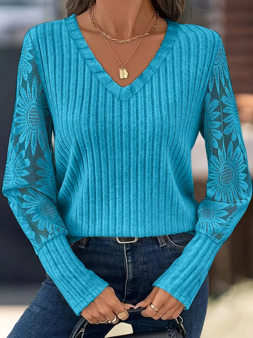 A woman wearing a turquoise blue ribbed knit sweater with floral lace sleeves, paired with dark blue jeans and gold necklaces.