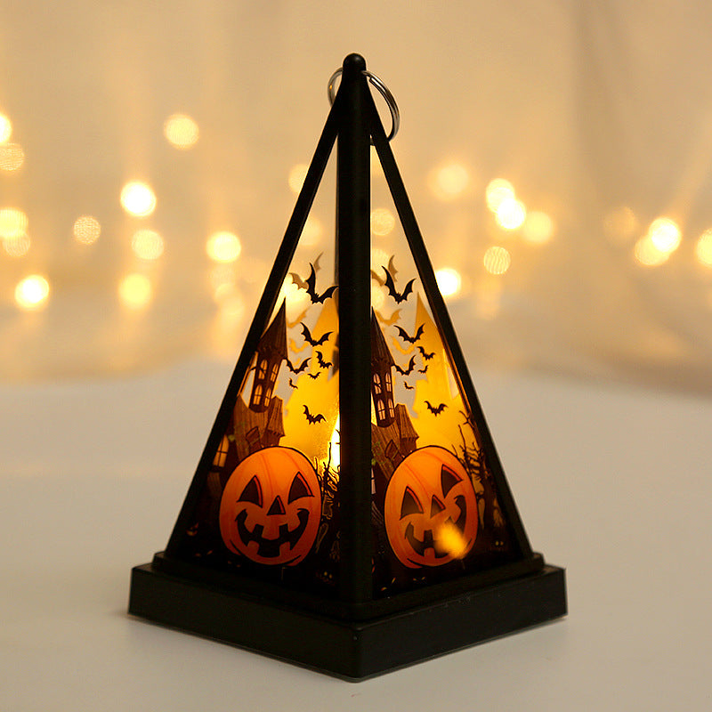 A black pyramid-shaped Halloween candle lantern with cutout silhouettes of a haunted house, bats, and jack-o'-lanterns, glowing with warm orange light against a softly blurred warm background.
