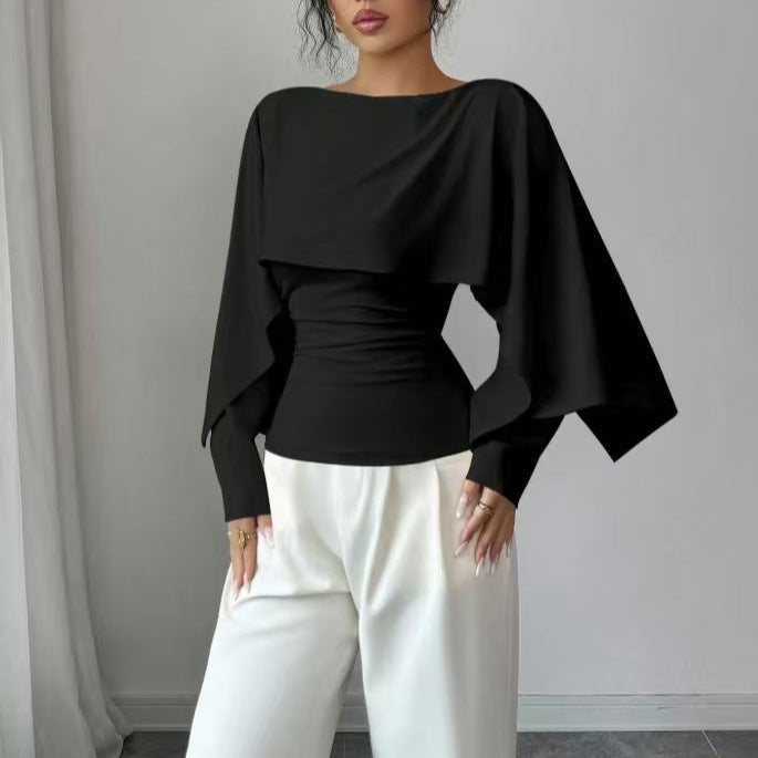 A woman wearing a black long-sleeve top with a wide boat neckline and dramatic draped cape sleeves, paired with white wide-leg pants, standing against a light gray wall.