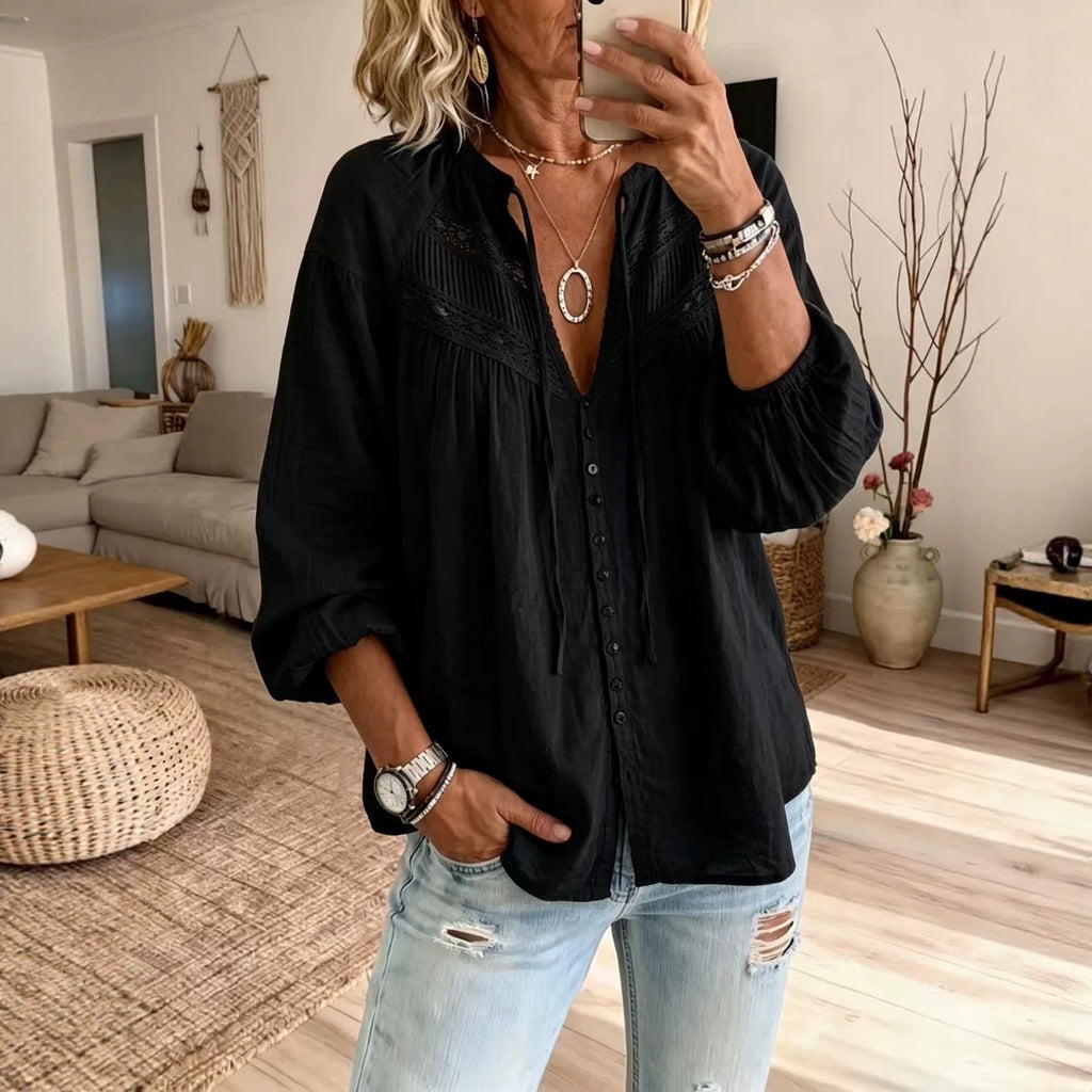 A woman takes a mirror selfie in a living room, wearing a black long-sleeve button-up blouse with gathered detailing at the chest and light-wash distressed jeans.