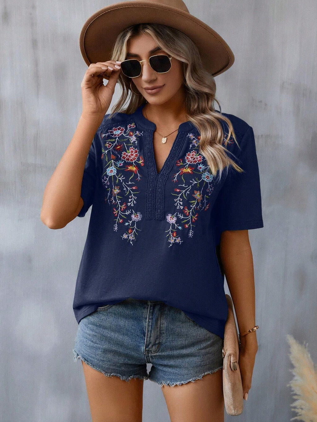 Woman wearing a navy blue embroidered top with floral designs, denim shorts, and a brown hat against a gray background.