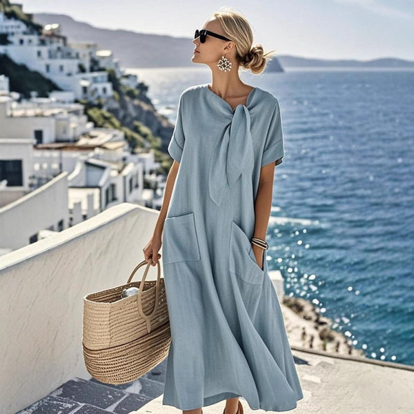Woman in a light blue dress standing on a balcony with a scenic view of the ocean and white buildings.