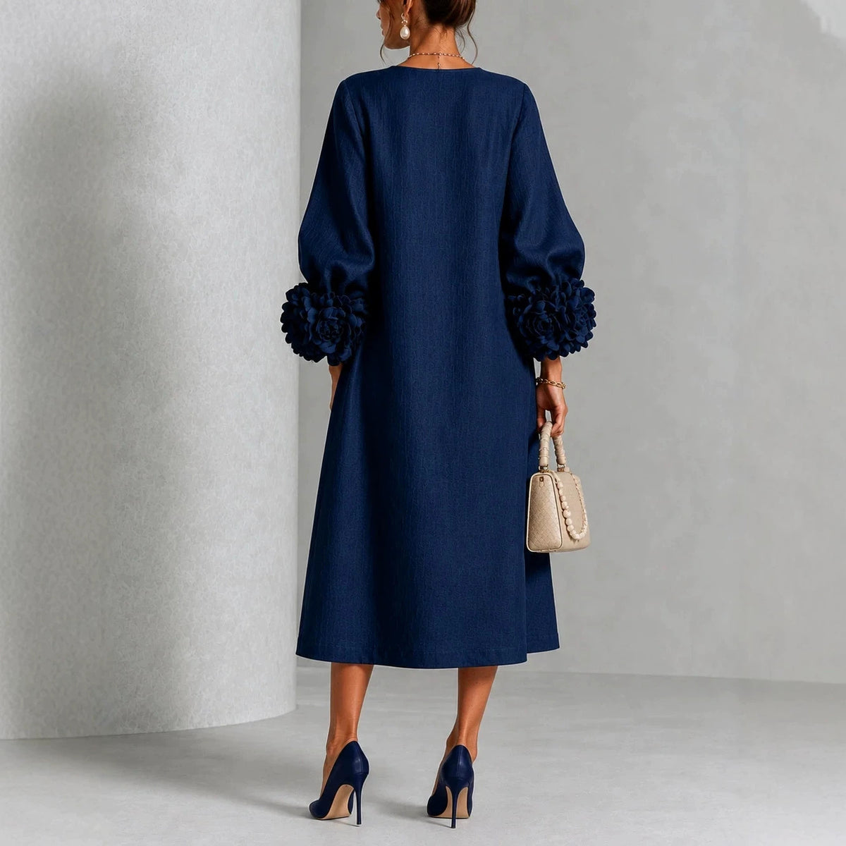 A woman shown from the back wearing a navy blue midi dress with voluminous long sleeves and ruffled cuffs, holding a small beige handbag and wearing navy heels against a light gray studio backdrop.