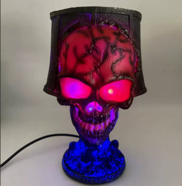 A skull-shaped lamp with a black textured shade and base, illuminated with red and blue LED lights, shown against a plain light gray background.