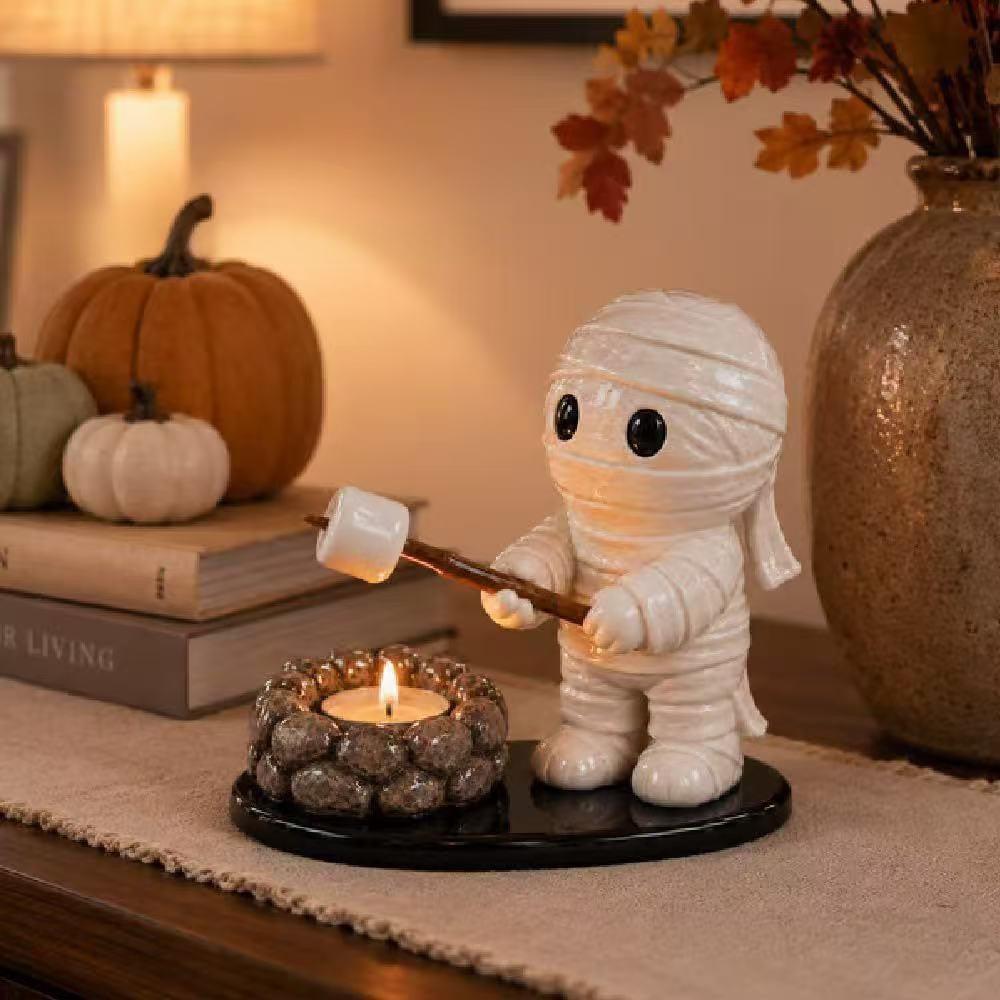 A mummy-shaped candle holder and tea light holder sits on a tabletop in a warm, autumnal setting. The white ceramic mummy figure holds a wooden stick with a marshmallow, standing beside a small lit candle in a stone-like ring base on a black oval platform.