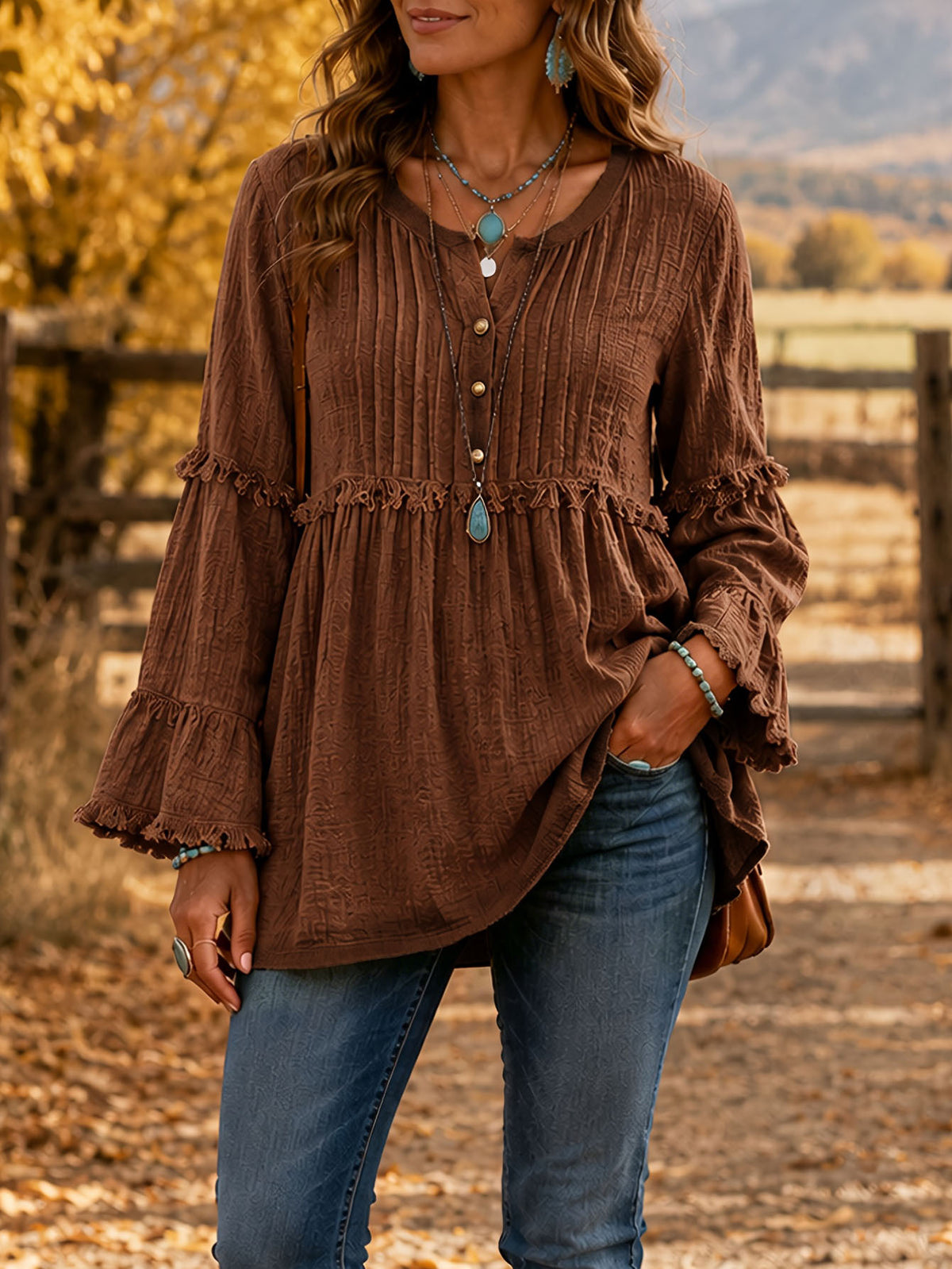 A woman wearing a brown long-sleeve blouse with a split V-neckline, button details, and ruffled cuffs, paired with blue jeans, standing outdoors in a rustic setting.