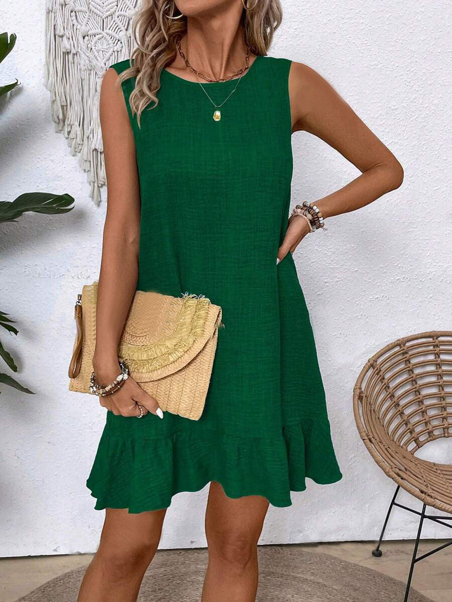 Woman wearing a green dress holding a beige handbag against a white wall.