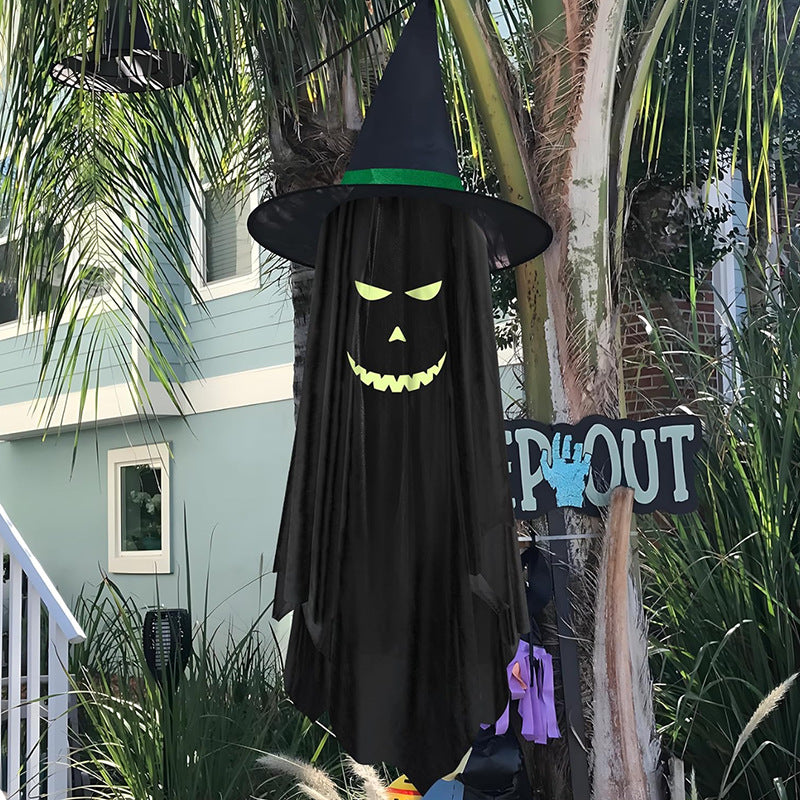 A hanging black ghost Halloween decoration with a witch hat and glowing neon-style face, displayed outdoors in front of a house with palm trees and a 'CROUT' sign nearby.