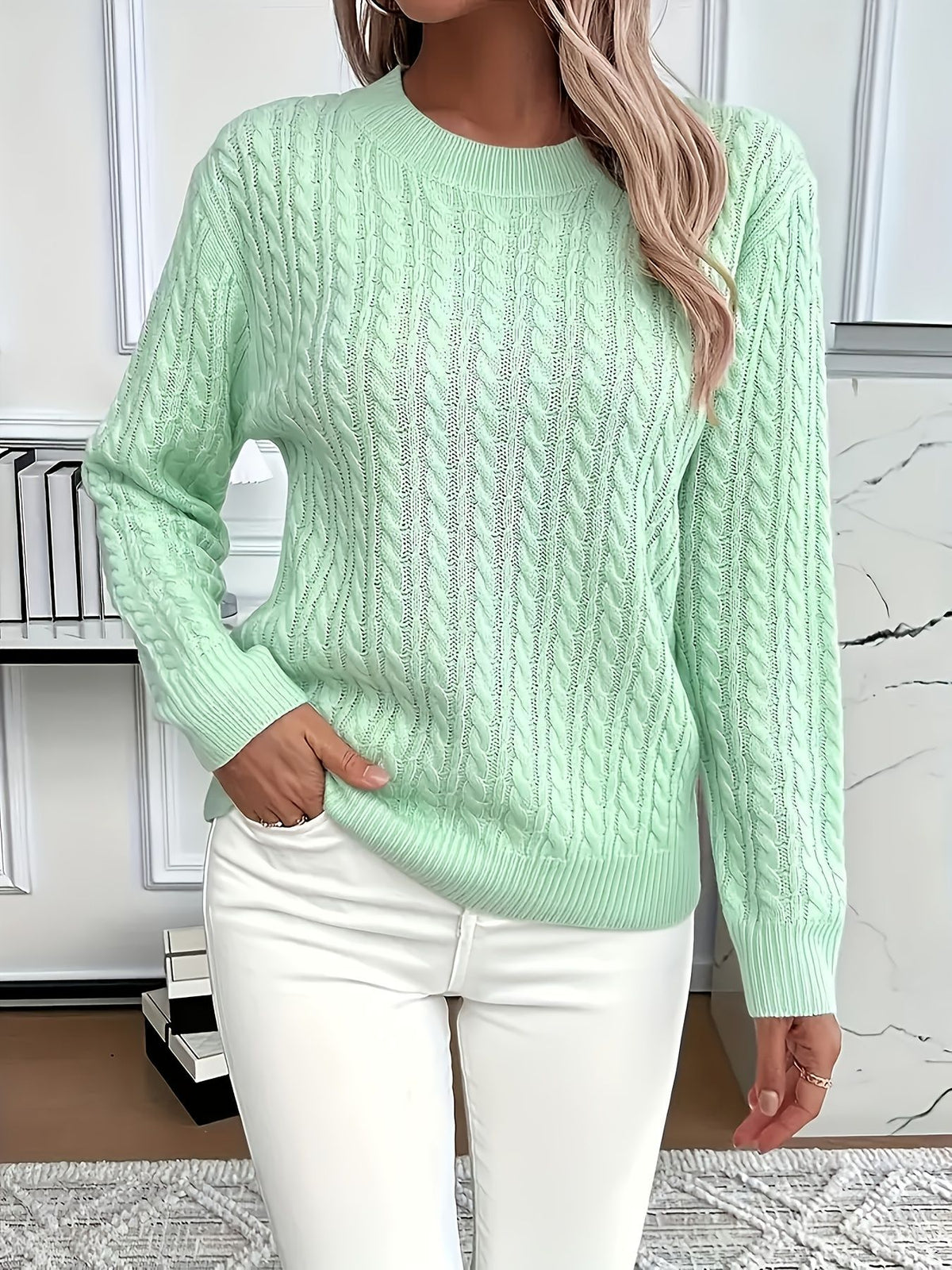 Person wearing a light green cable knit sweater and white pants in a room with a marble wall.
