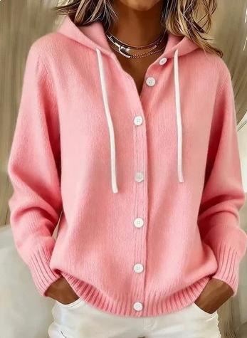A woman wearing a pink knit cardigan hoodie with white drawstrings and white buttons, paired with white pants and layered necklaces.