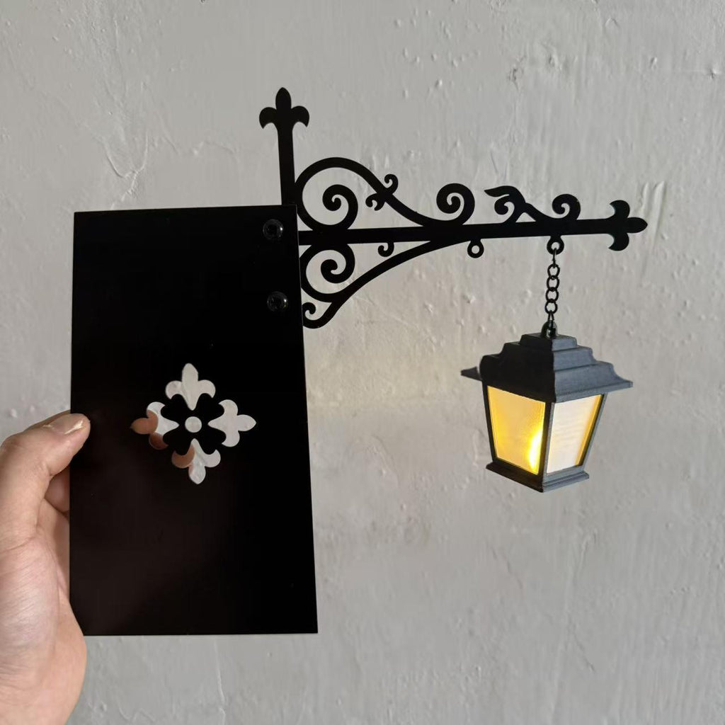 A decorative black wrought-iron style wall sconce with an ornate bracket and hanging lantern, shown mounted on a light gray wall. A hand holds a black card with a white floral cutout beside it for scale.