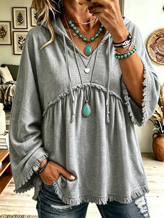 A woman wearing a loose gray hooded tunic top with drawstrings, fringe trim, and wide bell sleeves, paired with distressed blue jeans and layered turquoise jewelry in a home setting.