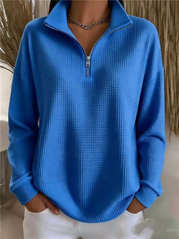 A person wearing a bright blue waffle-knit quarter-zip pullover sweater with a stand collar and white pants, shown from the neck down in an indoor setting.