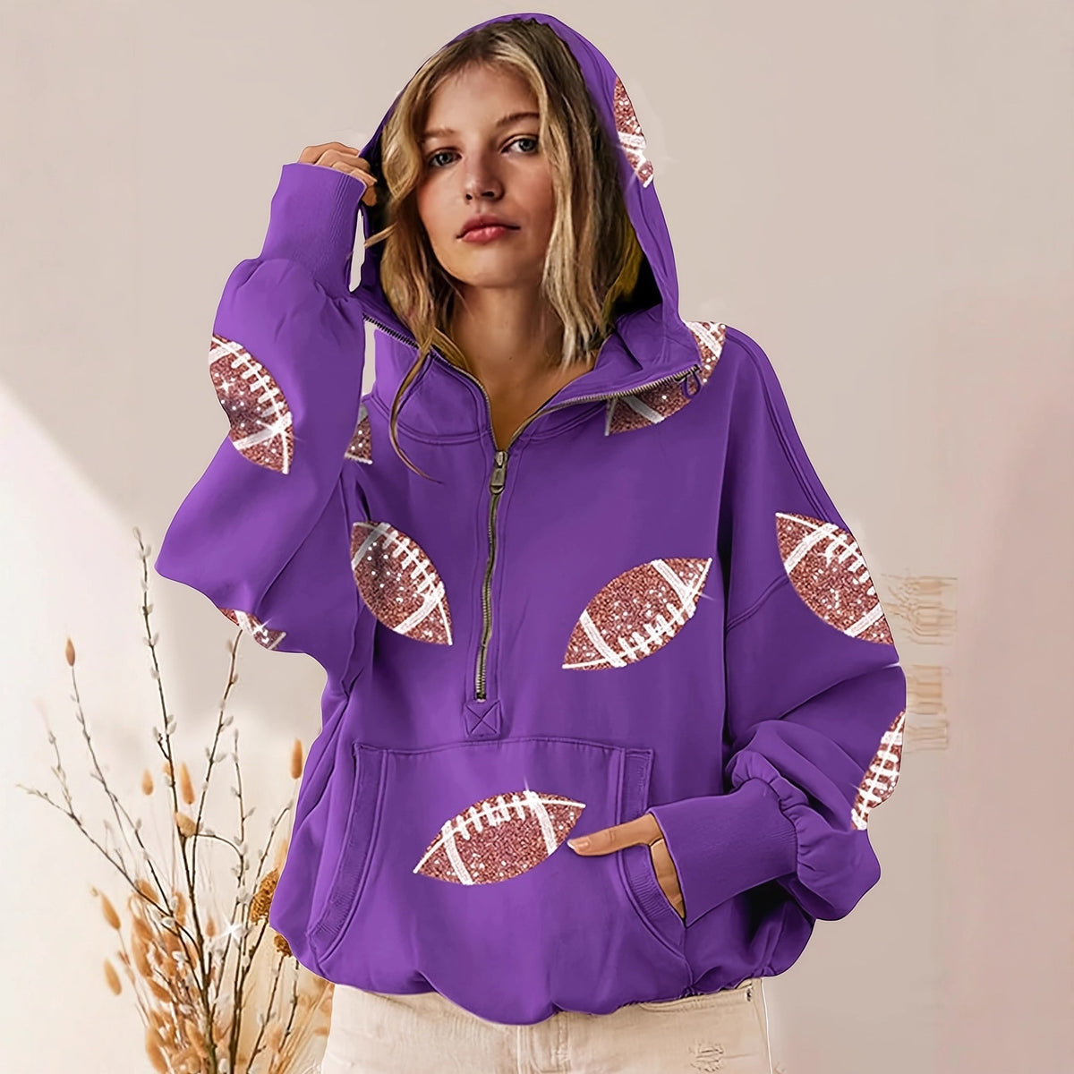 A woman wearing a purple half-zip hoodie with football-shaped sequin patches, posed against a warm beige background with dried grass decor.
