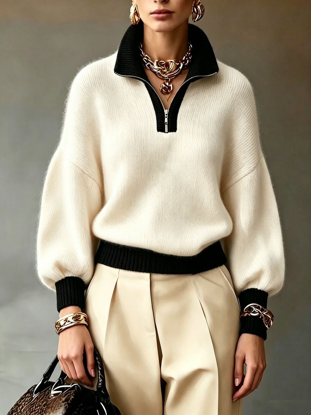 A woman models a cream-colored knit quarter-zip sweater with black ribbed trim at the collar, cuffs, and hem, paired with pleated cream trousers and gold jewelry.