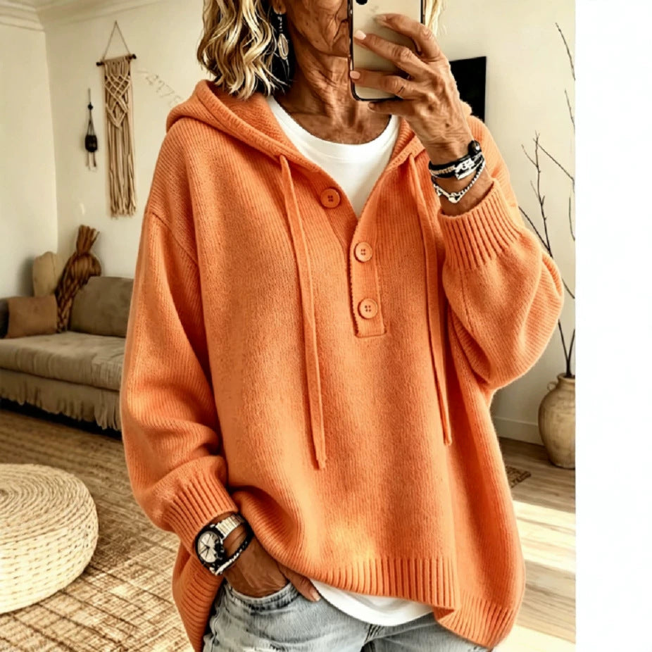 A woman takes a mirror selfie wearing an oversized orange knit hooded sweater with a button placket over a white top and light-wash jeans, in a cozy living room setting.