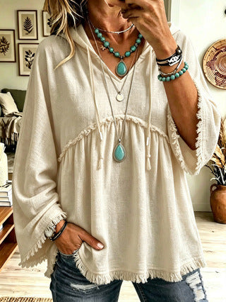 A woman wearing a cream-colored, loose-fitting bohemian-style tunic top with fringe details and drawstrings, paired with distressed blue jeans and turquoise jewelry.