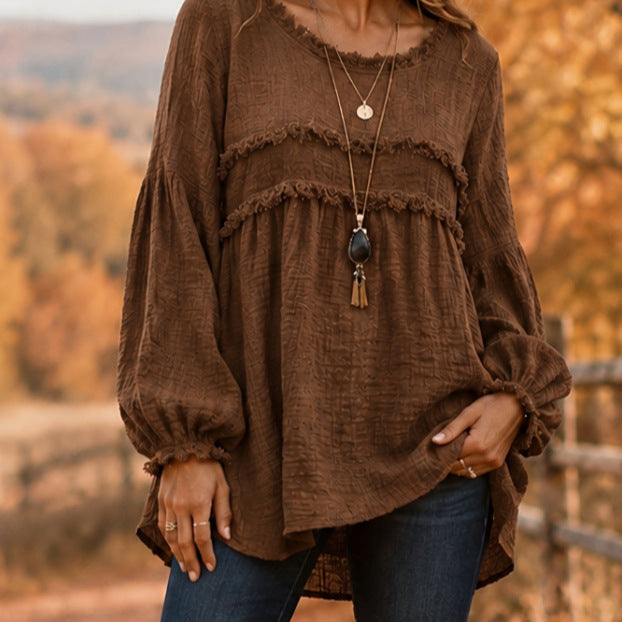 A woman wearing a brown crinkled cotton long-sleeve blouse with layered ruffle details and elasticized cuffs, paired with blue jeans, set against an outdoor autumn background.