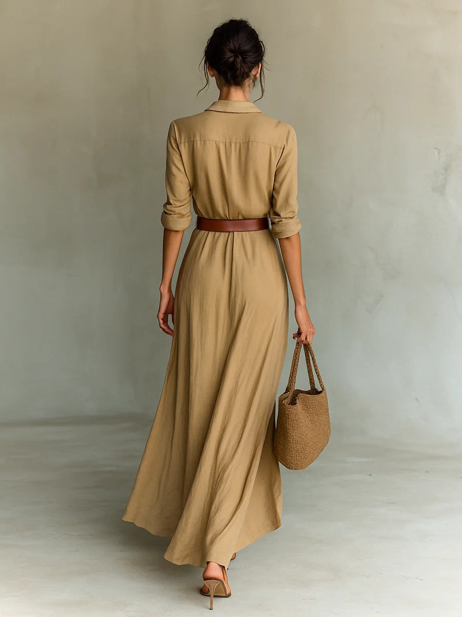Woman wearing a long beige dress with a brown belt and holding a matching handbag against a plain background.