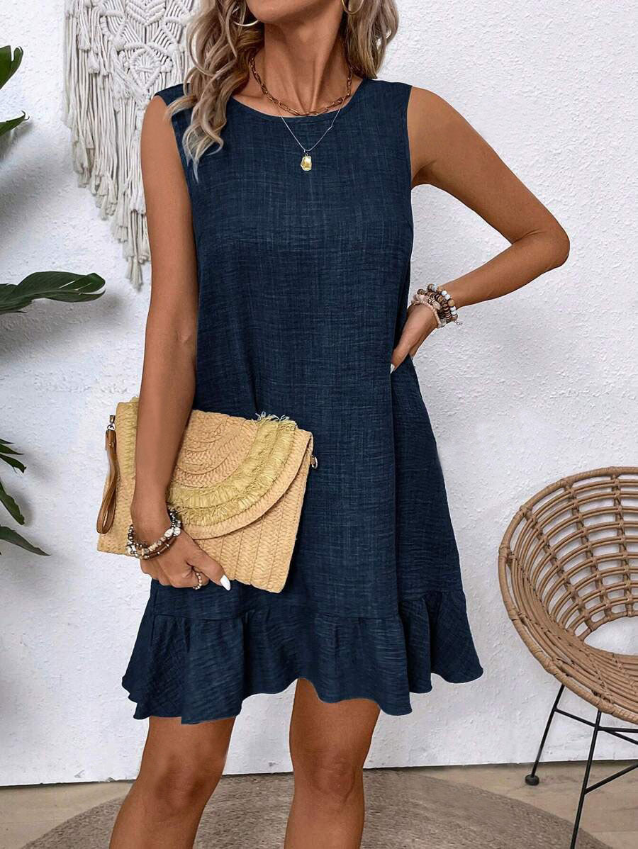 Woman wearing a navy blue dress holding a beige handbag against a white wall.