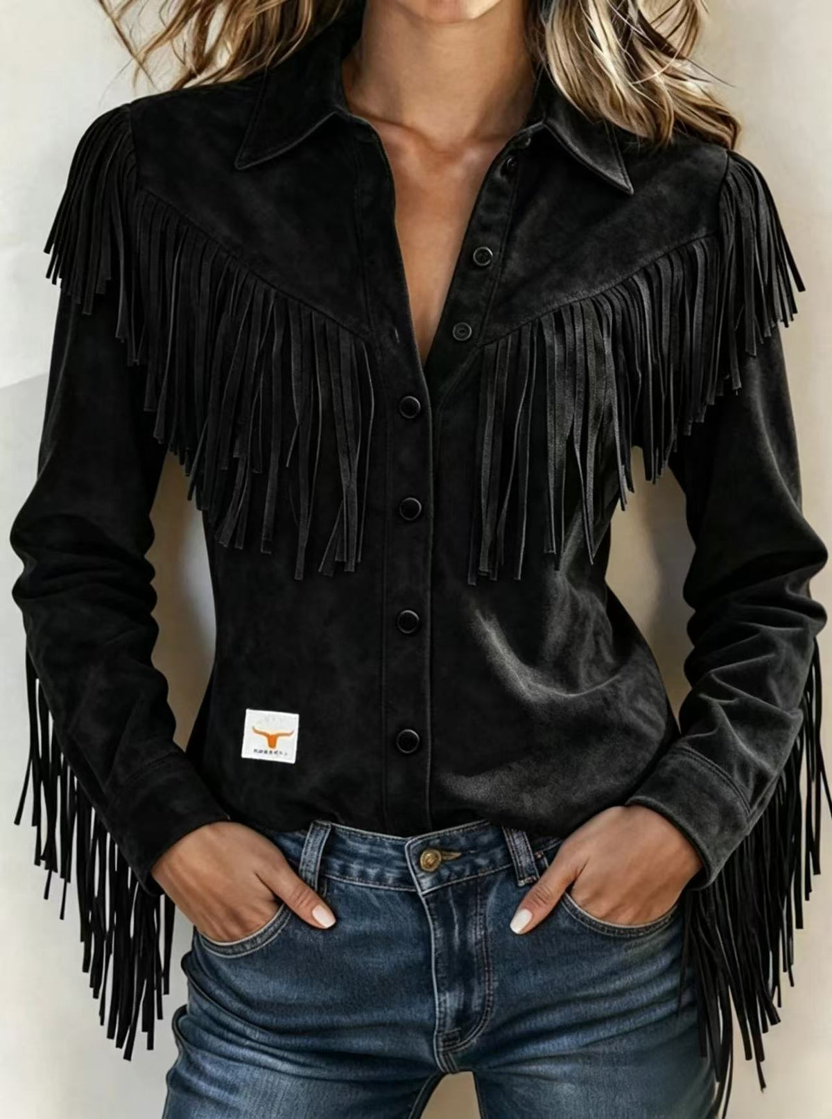 A woman wearing a black long-sleeve western-style shirt with heavy fringe detailing across the chest and shoulders, paired with blue jeans. A small white rectangular patch with an orange longhorn logo is visible on the lower left side of the shirt.