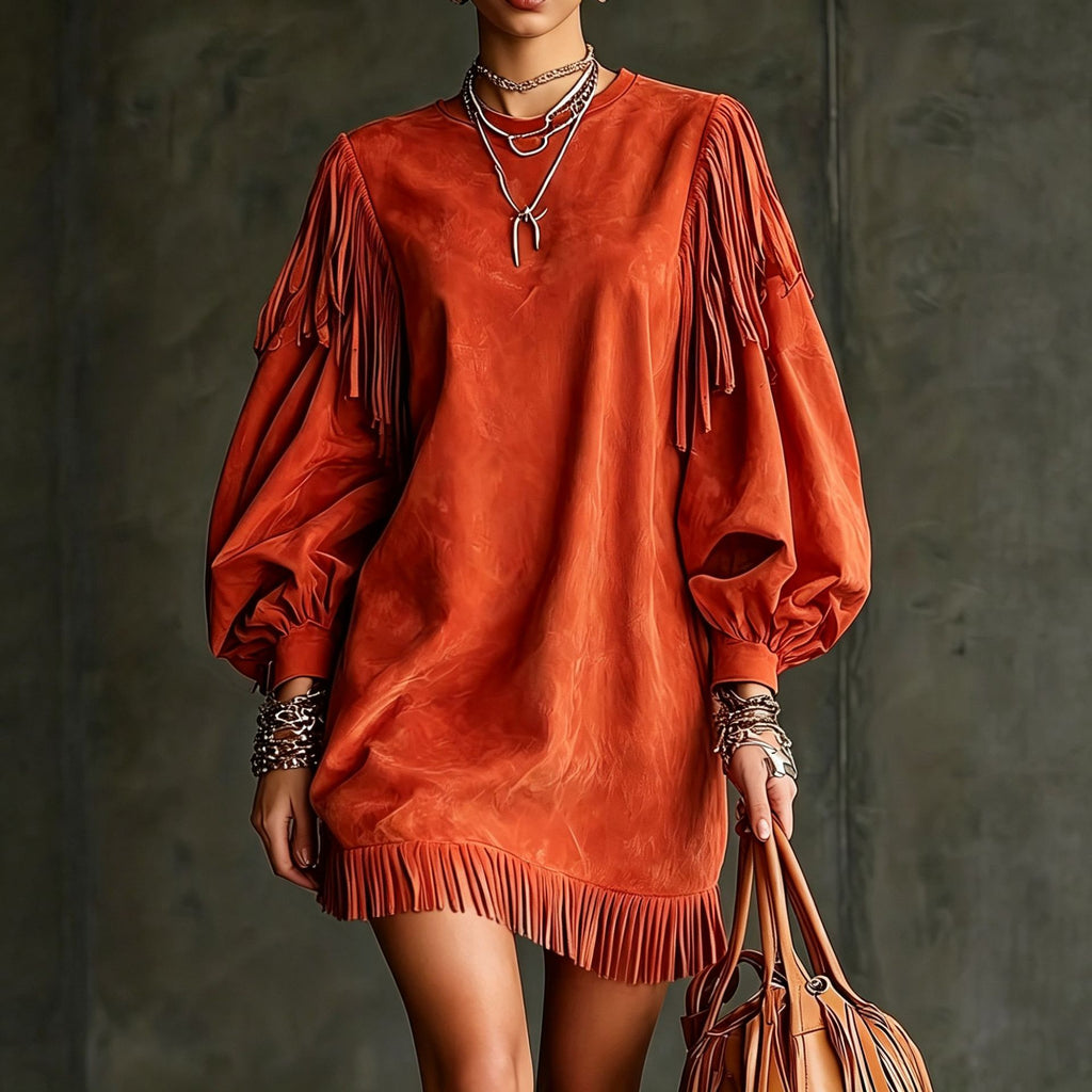 A model wears a burnt orange suede puff-sleeve mini dress with fringe detailing on the sleeves and hem, accessorized with layered silver necklaces and bracelets, holding a matching orange-brown fringe handbag against a dark textured studio background.