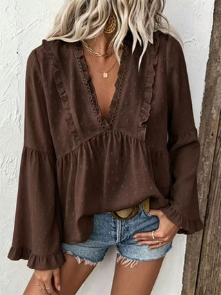 A woman wearing a dark brown long-sleeve blouse with ruffle trim and a deep V-neck, paired with denim shorts, standing against a white textured wall.