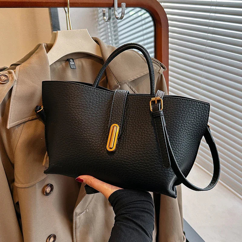 Black handbag held by a person with a beige trench coat in the background