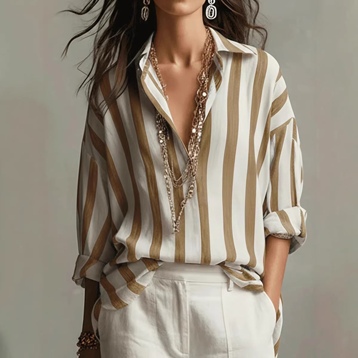 A woman wearing a loose white button-up shirt with vertical tan stripes, styled with layered gold necklaces and white trousers against a soft gray background.
