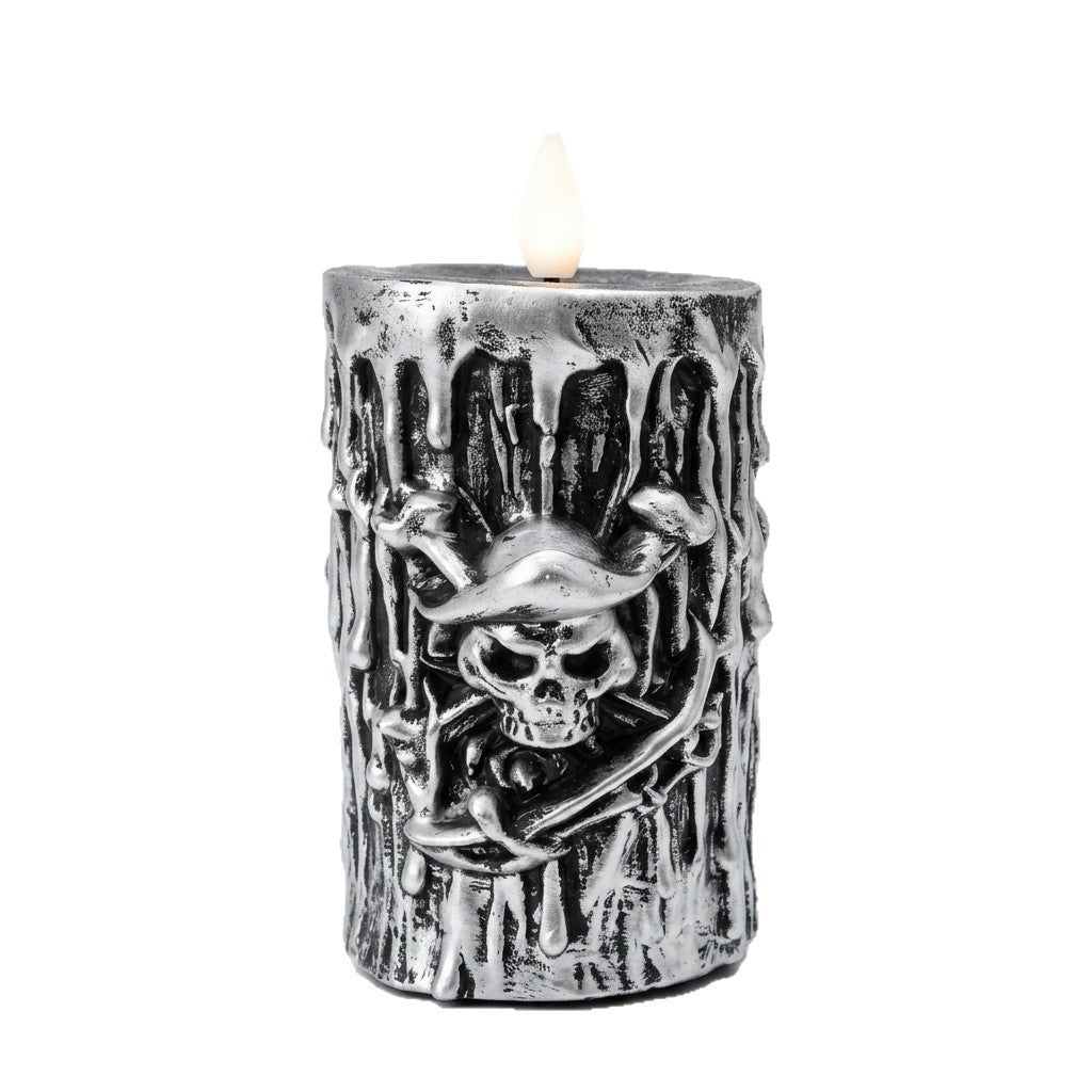 A silver-colored decorative pillar candle with a dripping wax effect and a raised skull and crossbones design on the front, shown lit against a plain white background.