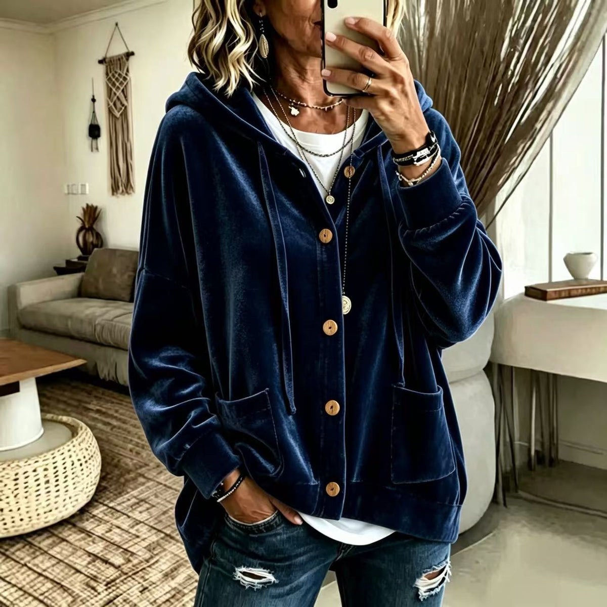 A woman takes a mirror selfie wearing a navy blue velvet zip-up hoodie with brown wooden buttons, layered over a white t-shirt and blue distressed jeans in a modern living room setting.