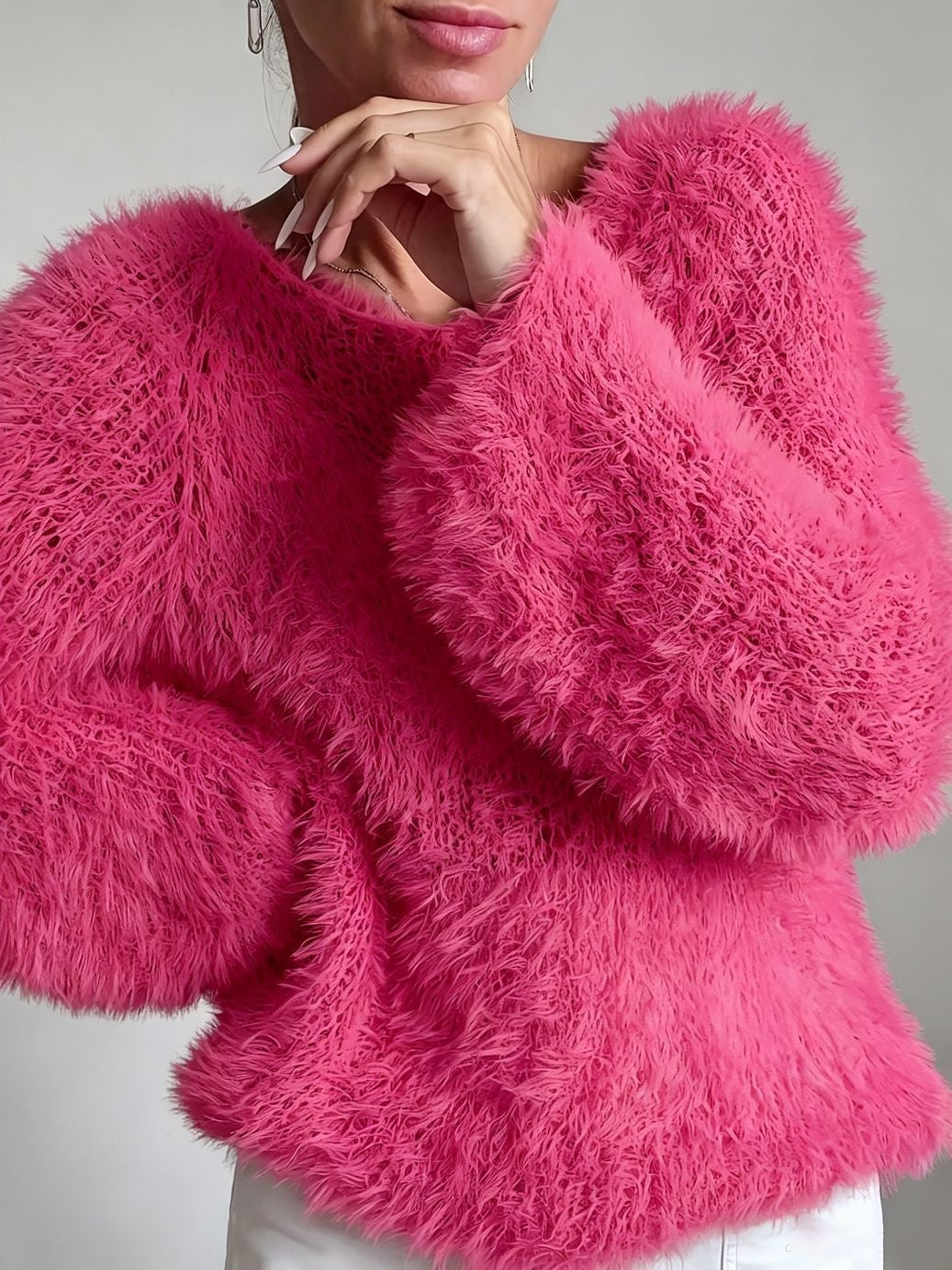 A close-up of a woman wearing a bright pink, voluminous faux fur sweater with dramatic puffed sleeves, paired with white pants against a neutral gray background.