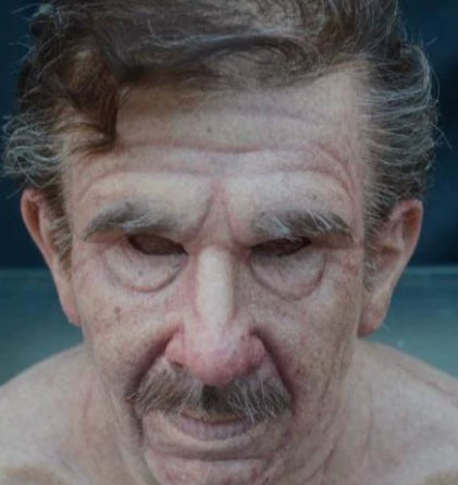 A close-up portrait of an older man with a mustache, showing detailed skin texture and wrinkles against a dark blue background.