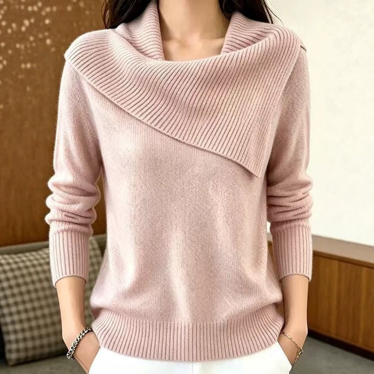 A woman wearing a light pink cowl-neck knit sweater with long sleeves and white pants, standing indoors against a neutral background.
