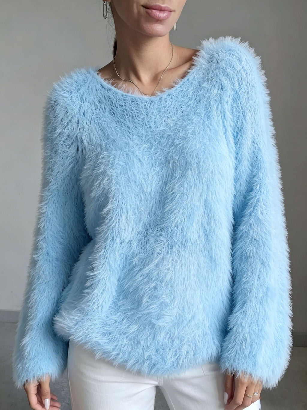 A woman wearing a light blue fuzzy long-sleeve sweater and white pants, shown from the mouth down against a plain gray wall.