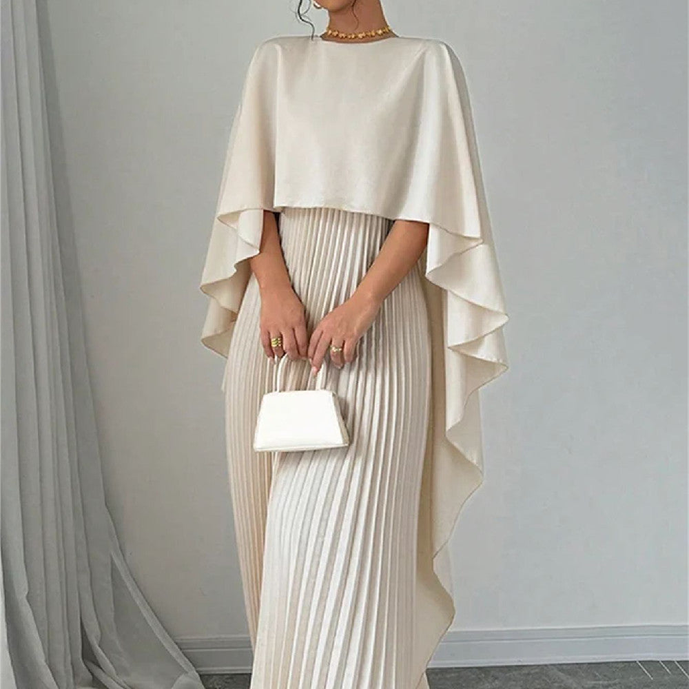 Woman wearing a beige cape over a pleated dress, holding a matching handbag against a gray background.