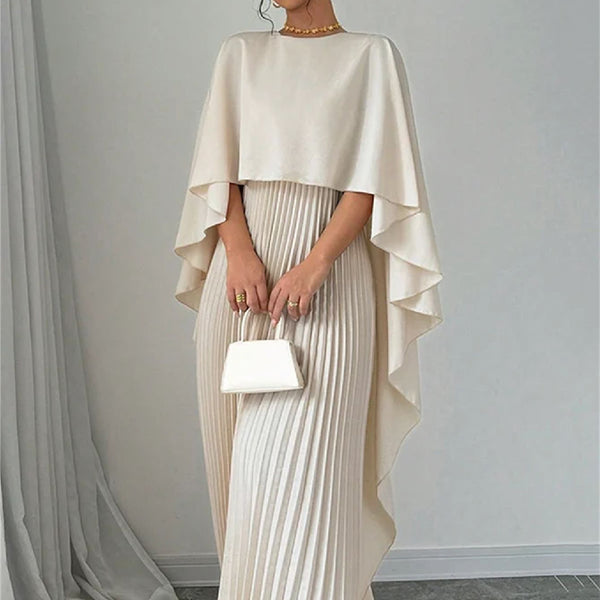 Woman wearing a beige cape over a pleated dress, holding a matching handbag against a gray background.