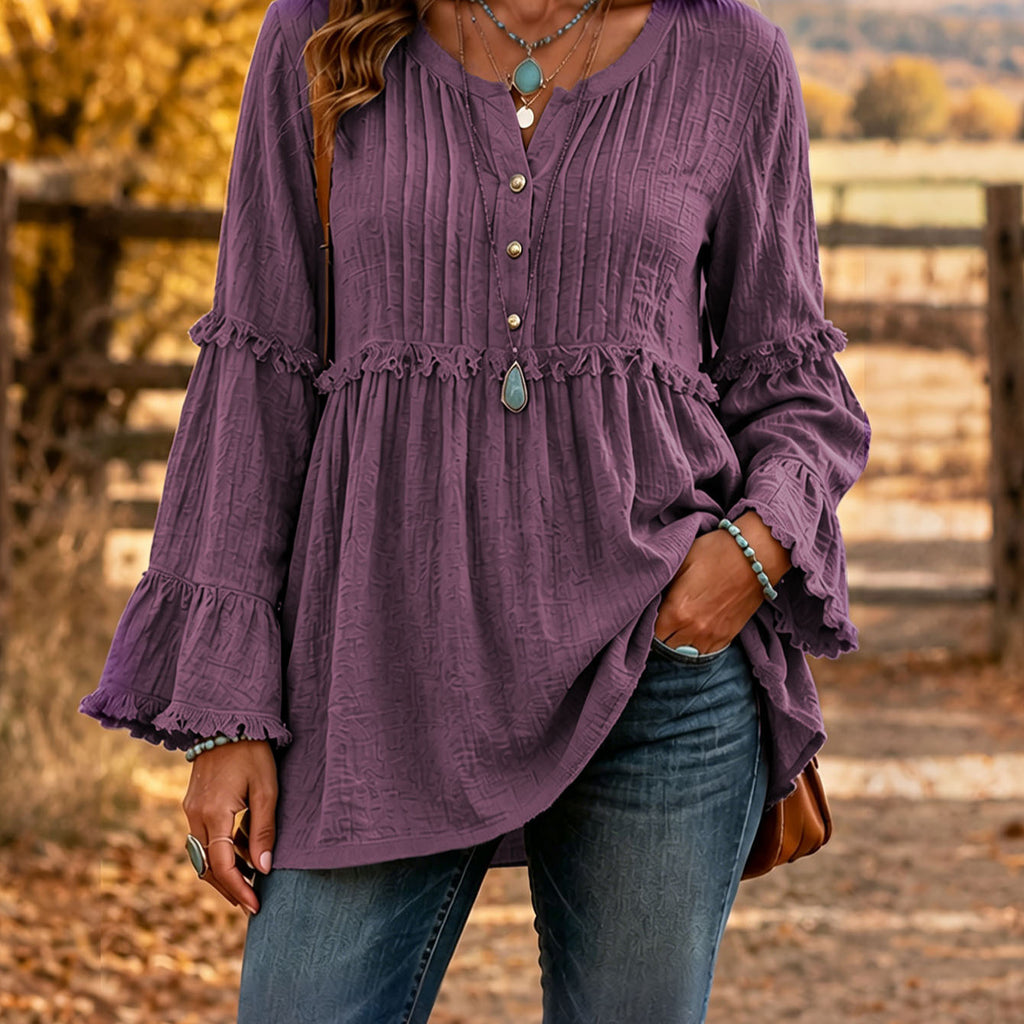 A woman wearing a textured purple long-sleeve blouse with ruffle details and a button-down front, paired with blue jeans, standing outdoors in front of a rustic wooden fence.