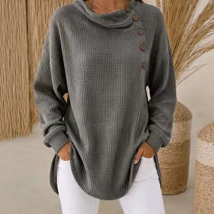 A woman wearing a gray waffle-knit cowl-neck sweater with brown buttons on the side, paired with white pants, standing in a styled indoor setting with woven decor.